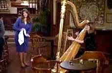 Two women in a bar, one standing holding a bar towel speaking to another seated at a café table playing her full-size orchestra harp.